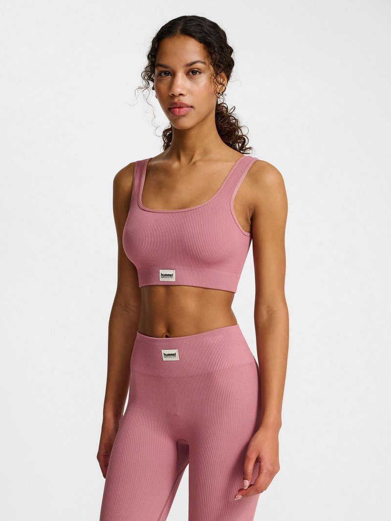 Kvinde i pink ribbet crop top og højtaljede leggings stående imod en ensfarvet hvid baggrund, vendt fremad med afslappet udtryk.