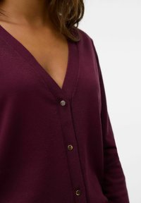 Cardigan bordeaux con scollatura a V profonda, morbida texture in maglia e cinque bottoni metallici sul davanti, con maniche lunghe e una forma adatta.