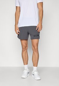 Halli spordilühikesed hallid, millel on külgedel must "BORG" logo, koos valge lühikeste varrukatega särgi ja valgete spordijalatsitega.