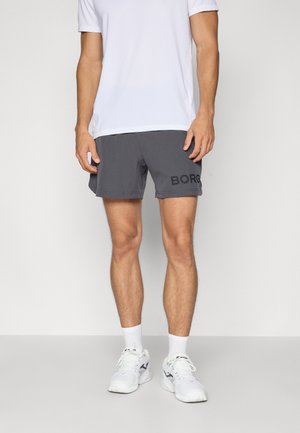 Moški, ki nosi bele športne copate, bele nogavice, sive športne kratke hlače z napisom "BORG" in belo majico s kratkimi rokavi, stoji ob belem ozadju.