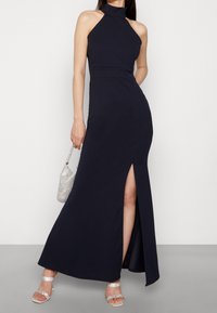 Vestido maxi azul marino con cuello halter, abertura lateral, tela suave, cuerpo ajustado, combinado con un bolso de cuentas plateadas y sandalias con tiras.