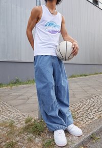 Biała bezrękawnik z grafiką "Havers", luźne jasnoniebieskie dżinsy, białe sneakersy z czerwonymi akcentami, trzymane piłka koszykowa.