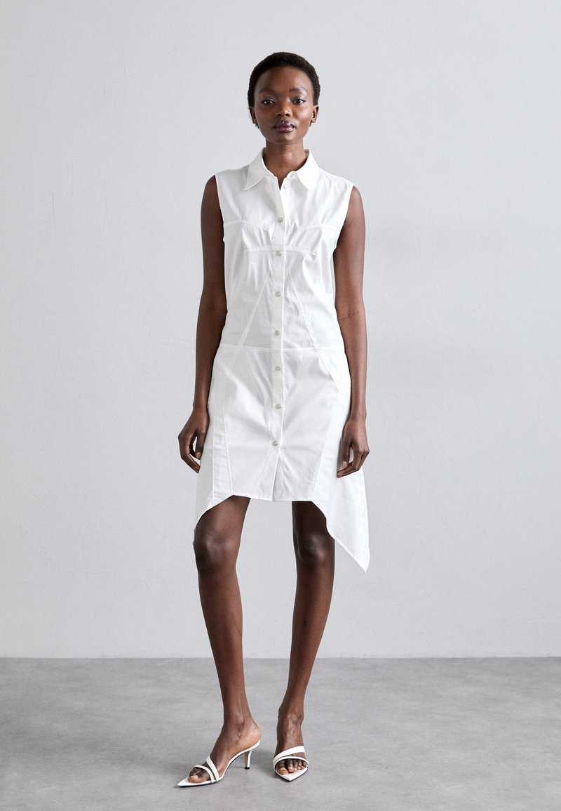 Fehér ujjatlan ingruha gombos elülső résszel, aszimmetrikus szegéllyel és testhez álló fazonnal; fehér nyitott orrú magassarkúval párosítva.