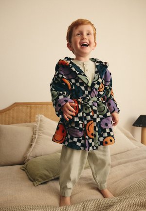 Smiling red-haired child standing on bed wearing patterned fleece robe with colorful smiley faces and checkered design over cream pajamas.