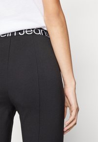 Schwarze Leggings mit einem elastischen Bund mit Markenlogo. Der Stoff wirkt glatt und hat ein eng anliegendes Design, dargestellt aus einer Seitenansicht.