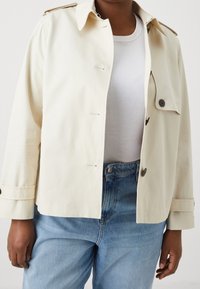 Osoba w jasnobeżowej kurtce na białej prążkowanej bluzce i jasnoniebieskich wysokich spodniach dżinsowych, stojąca na jednolitym tle.