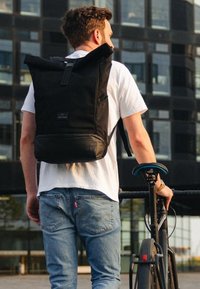 Egy fehér pólót és kék farmert viselő férfi fekete kerékpárt tart, és egy nagy fekete hátizsák van a hátán, miközben egy modern üvegből készült épület előtt áll.