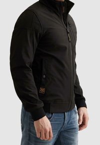 Homme portant une veste noire à zip avec un col montant et un jean bleu, debout devant un fond uni clair.