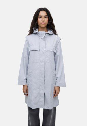 Mujer con cabello largo y oscuro, vistiendo un impermeable con capucha gris claro con bolsillos de solapa y pantalones gris oscuro, de pie sobre un fondo liso.