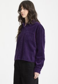 Lila kordflodds skjorta med knappframsida, krage och en enda bröstficka. Har ribbad textur och långa ärmar. Svarta byxor bärs.