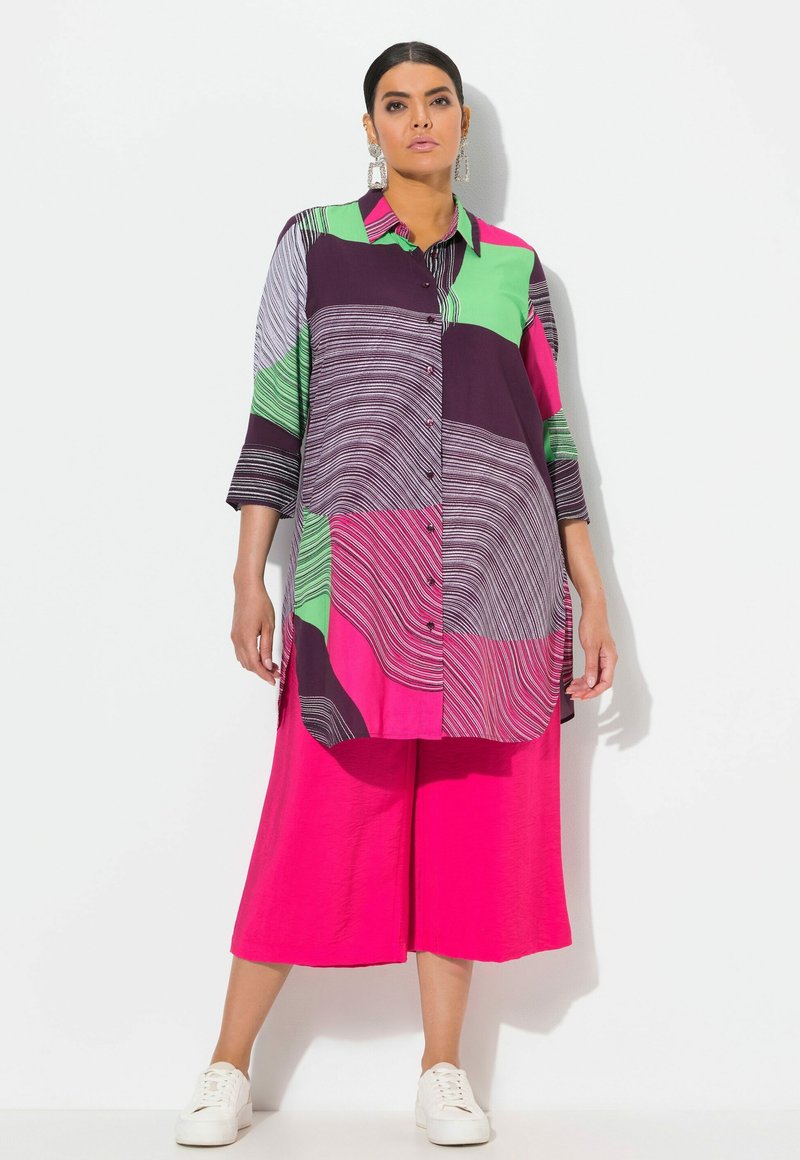 Camicia colorata in viola, verde e rosa con motivi ondulati, maniche lunghe e chiusura con bottoni, abbinata a pantaloni larghi rosa brillante.