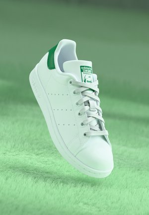 Witte Adidas Stan Smith sneaker met een groen hielstuk en logo op de tong, zwevend boven een lichtgroene getextureerde ondergrond.