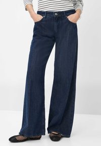 Weite, dunkelblaue Jeans mit hoher Taille, mit einem Knopfverschluss, Fronttaschen und einer glatten Denim-Oberfläche. Kombiniert mit schwarzen Mesh-Ballerinas.