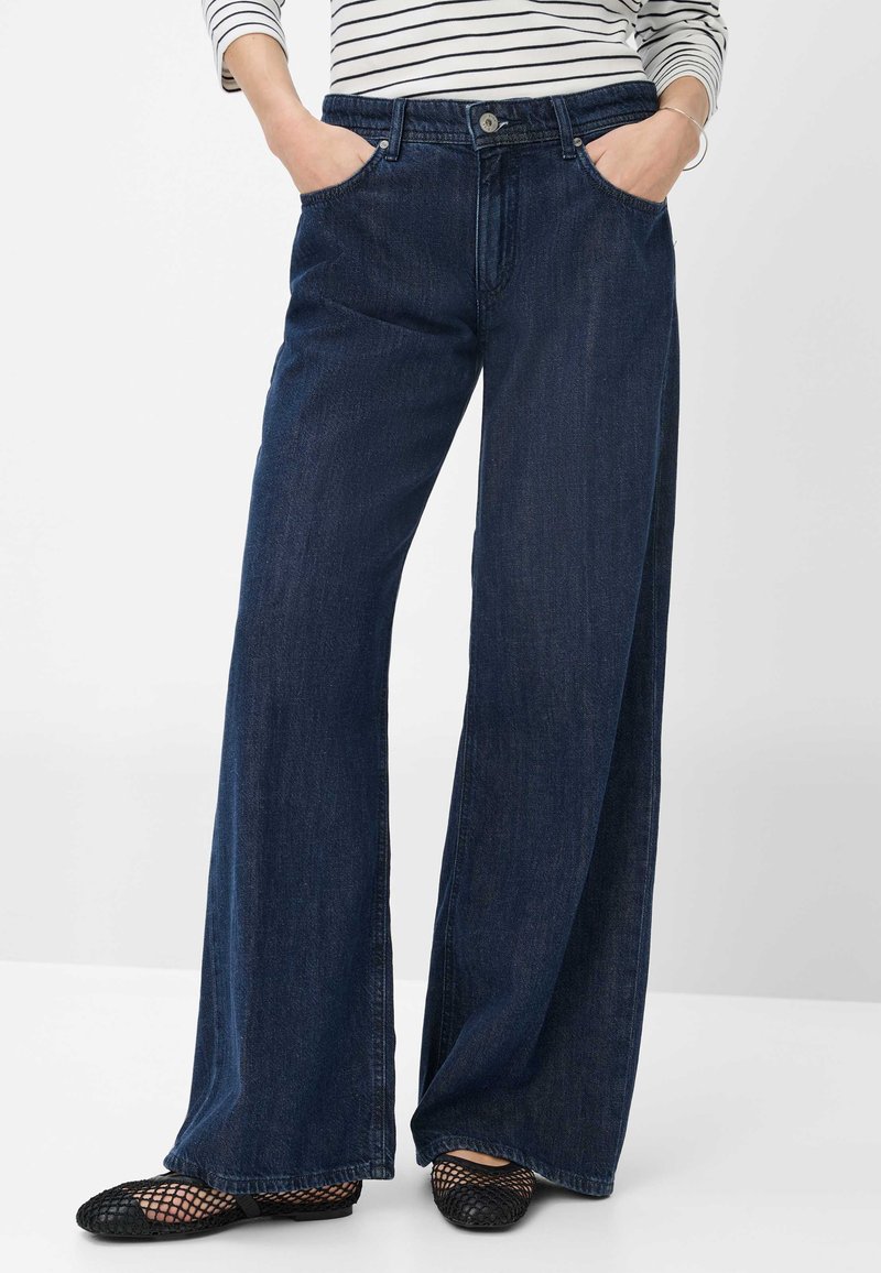 Weite, dunkelblaue Jeans mit hoher Taille, mit einem Knopfverschluss, Fronttaschen und einer glatten Denim-Oberfläche. Kombiniert mit schwarzen Mesh-Ballerinas.