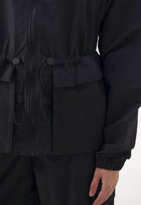 Chubasquero negro con cintura ajustada, cierre frontal con cremallera, puños elásticos y bolsillos funcionales. Fabricado con tela ligera y texturizada.