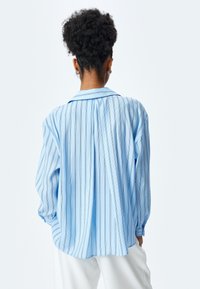 Camisa de rayas azul claro con mangas largas y sueltas, un collar clásico y un dorso ligeramente fruncido, que presenta finas líneas verticales en un color contrastante.