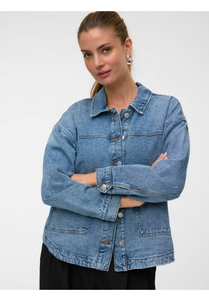 Femme aux cheveux attachés, portant des boucles d'oreilles en anneau et une veste en jean bleue, bras croisés, regardant légèrement souriante vers la caméra.