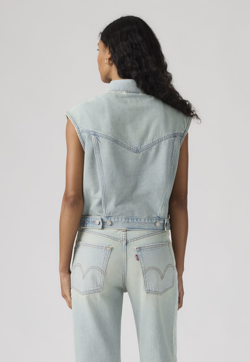 Femme aux longs cheveux bouclés portant une veste en jean sans manches bleu clair et un jean assorti, vue de dos sur un fond uni.