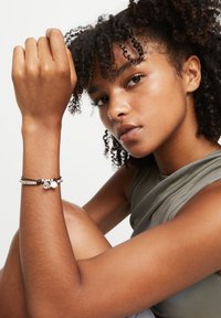 El brazalete presenta una mezcla de cuentas plateadas, cordón negro y detalles blancos. La modelo viste una camiseta sin mangas gris, con el cabello rizado enmarcando el rostro.