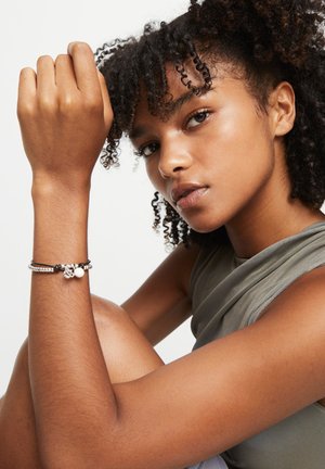 Le bracelet présente un mélange de perles argentées, de cordon noir et d'accents blancs. Le modèle porte un haut gris sans manches, avec des cheveux bouclés encadrant le visage.