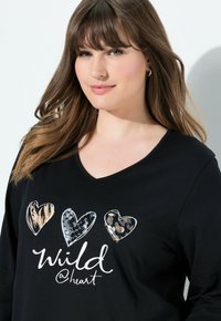 Schwarzes Langarmshirt mit drei Herzgrafiken in verschiedenen Tiermustern und dem Text "Wild @heart" in weißer, geschwungener Schrift.