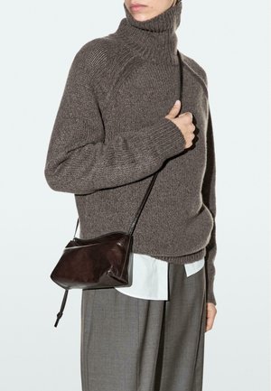 Donna che indossa un maglione marrone a collo alto con lavorazione a maglia grossa, pantaloni grigi, camicia bianca e una piccola borsa a tracolla in pelle marrone scuro.