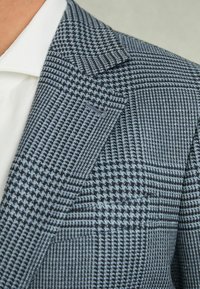 Blazer cu tipar houndstooth albastru și gri, din țesătură texturată, cu rever tăiat și un buzunar pe piept, purtat peste o cămașă albă.