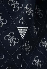 Marineblauer Stoff mit weißem, ineinandergreifendem "GS"-Muster und einem weißen, dreieckigen Guess U.S.A.-Logoaufnäher mit der Aufschrift "washed jeans".