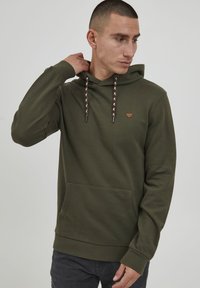 Maslinasto zeleni dukserica s kenguruznim džepom, rebrastim rukavima i kapuljačom koja ima šare na vezicama. Mali trokutasti logo na prsima.