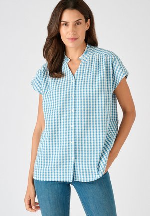 Mujer con blusa de manga corta con botones y cuello con volantes en cuadros azul y blanco y vaqueros azules, de pie contra un fondo blanco.