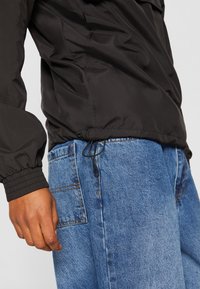 Sort nylonjakke med elastiske manchetter, snøret kant, parret med blå denimjeans med en sidelomme og subtile syninger.