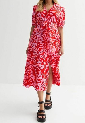 Frau trägt ein rotes und rosa geblümtes Midikleid mit kurzen Ärmeln und schwarzen Plateau-Sandalen, steht vor einem schlichten weißen Hintergrund.