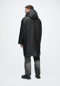 Jachetă neagră, impermeabilă, cu glugă și design lung, având o textură netedă. Logo roșu, notabil, pe mânecă. Purtați cu blugi gri deschis.