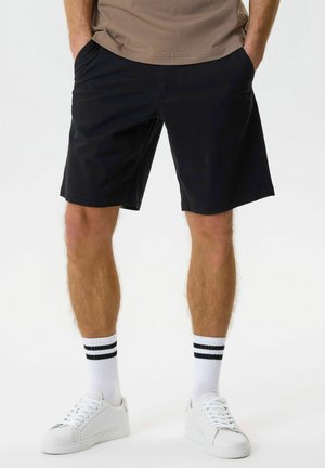 Férfi fekete rövidnadrágban, fehér sportcipőben és fehér bokazokniban, amelyen két fekete csík fut, zsebébe tett kézzel áll fehér háttér előtt.