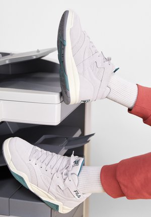 Sneakers in suede grigio chiaro con lacci bianchi, dettagli in teal e suole in gomma a contrasto; esposti su una superficie grigia di una stampante.