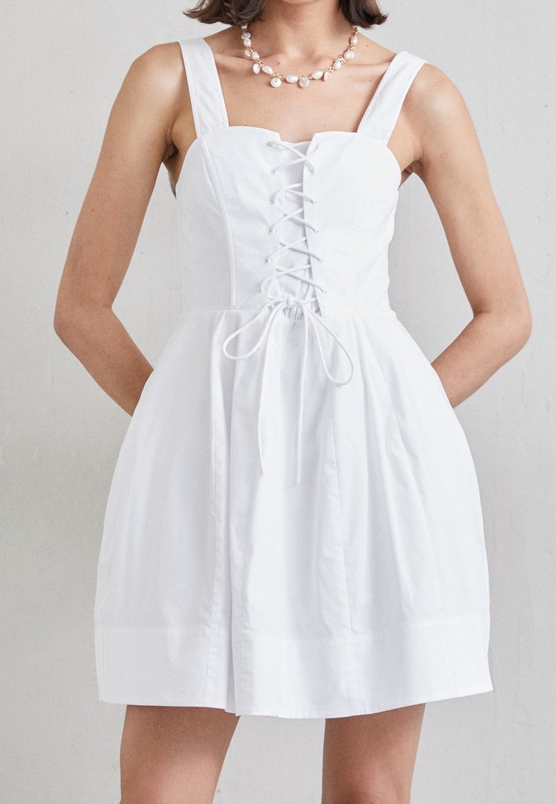 Femme portant une robe blanche sans manches avec un détail à lacets à l'avant et des bretelles larges, accessoirisée d’un collier de perles blanc.