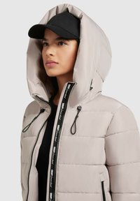 Veste rembourrée beige clair avec une grande capuche, deux poches zippées, bande de fermeture éclair noire avec logo, portée avec une casquette noire.
