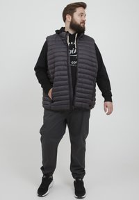Vadderad, ärmlös väst i mörkgrått, med en texturerad, quiltad design. Kombineras med en svart hoodies och grå byxor, bärs med svarta sneakers.