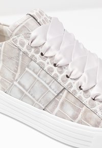 Teniska z texturované světle šedé kůže s krokodýlím motivem, má širokou saténovou tkaničku v jemně růžové barvě a masivní bílou gumovou podrážku.