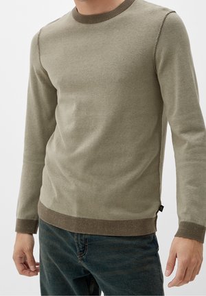 Mann trägt olivgrünen Pullover mit Rundhalsausschnitt, gerippten Bündchen und Saum, kombiniert mit dunkelblauen Jeans, steht vor einem schlichten Hintergrund.