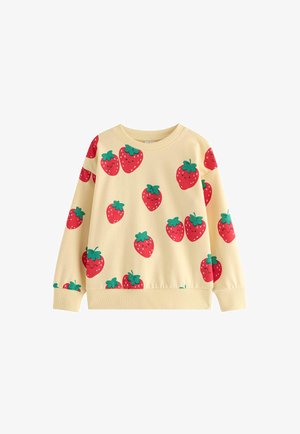 Ung pige iført en gul sweatshirt med jordbærprint, viddebenede jeans, røde sko og en lille lyserød crossbody-taske på hvid baggrund.