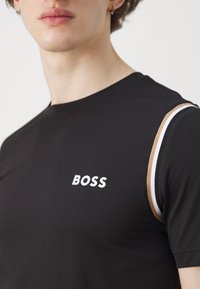 Camiseta negra ajustada con tejido texturizado, que presenta un logo blanco de "BOSS" en el pecho y una franja de acento en tonos beige y blanco en el hombro.