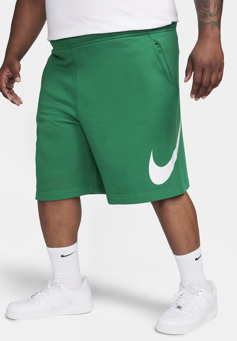 Mężczyzna w zielonych szortach Nike z dużym białym logo, białych skarpetach Nike i białych sneakersach Nike na jednolitym tle.