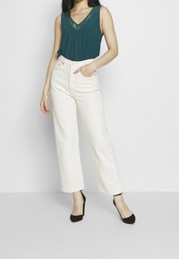Top senza maniche color teal con scollatura in pizzo, abbinato a pantaloni bianchi a gamba larga con vita alta e scarpe col tacco nere. Design semplice, pochi dettagli.