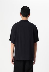 Camisa negra de manga corta hecha de tejido ligero; presenta un cuello y un diseño de corte recto con un dobladillo trasero ligeramente más largo.