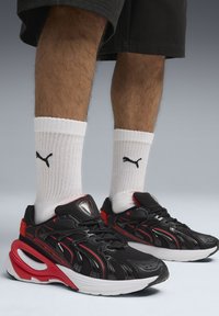 Baskets athlétiques noires et rouges avec une tige texturée, une semelle incurvée distinctive et un logo bien visible. Portées avec des chaussettes blanches côtelées.