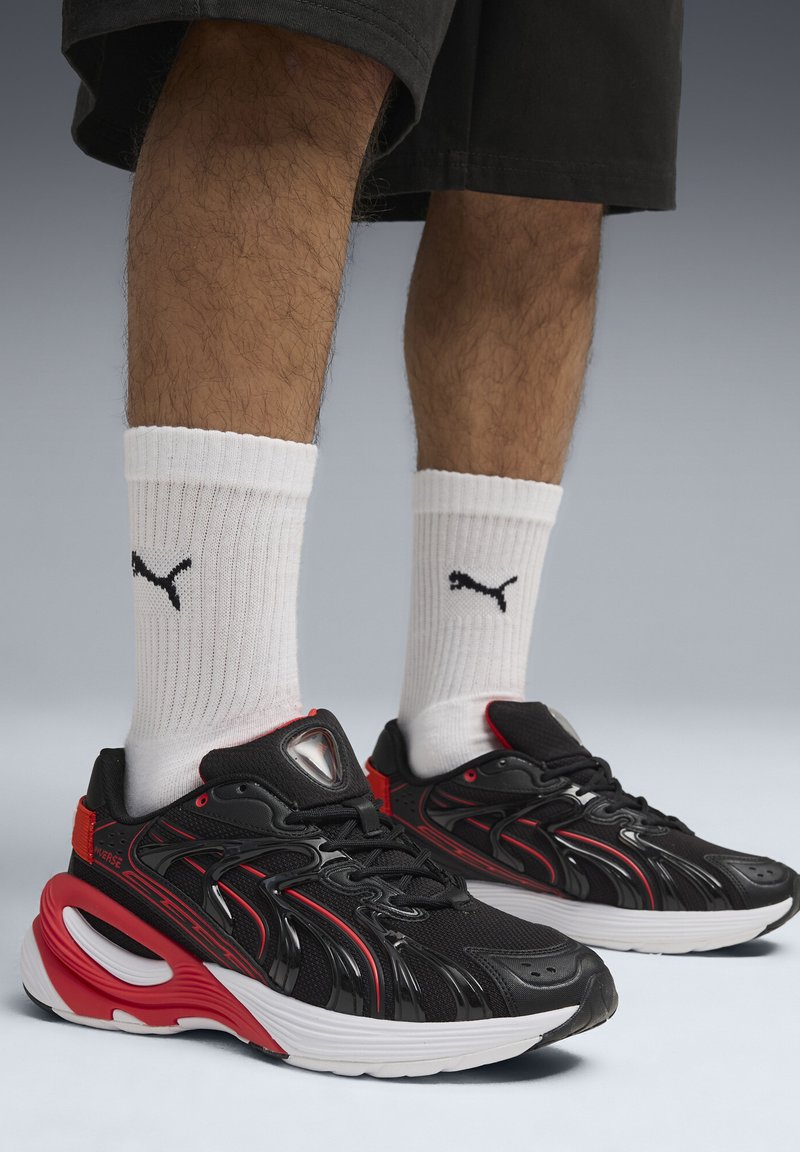 Baskets athlétiques noires et rouges avec une tige texturée, une semelle incurvée distinctive et un logo bien visible. Portées avec des chaussettes blanches côtelées.