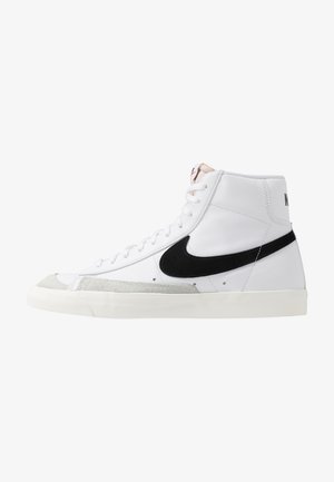 White high-top sneakers with a black swoosh, textured gray toe cap, and flat white sole. Features six eyelets and stitched accents.