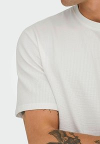 Hvid kortærmet t-shirt med et tekstureret mønster, rund halsudskæring og let rullet ærmeafslutning. Synlig tatovering på armen.