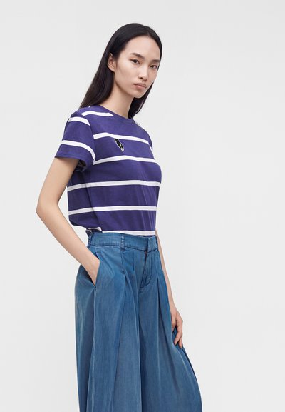 Jeune femme aux longs cheveux noirs portant un t-shirt rayé violet et blanc et un pantalon large bleu, les mains dans les poches.